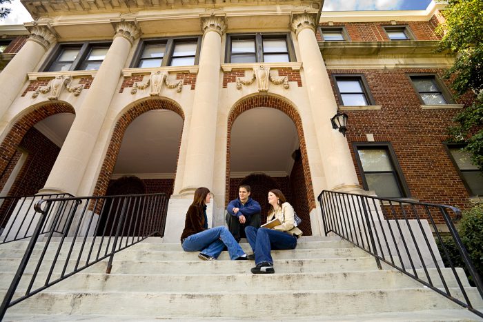 Three people sitting and talking on the steps of a large building.