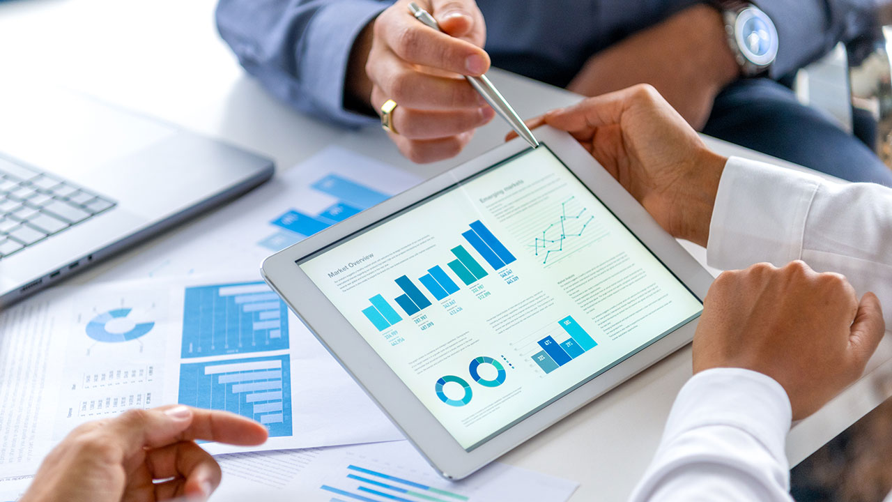 Close up of three people looking at financial data with graphs and charts. All their hands can be seen and one person is pointing with a pen. There is paperwork on the desk showing more finance information.
